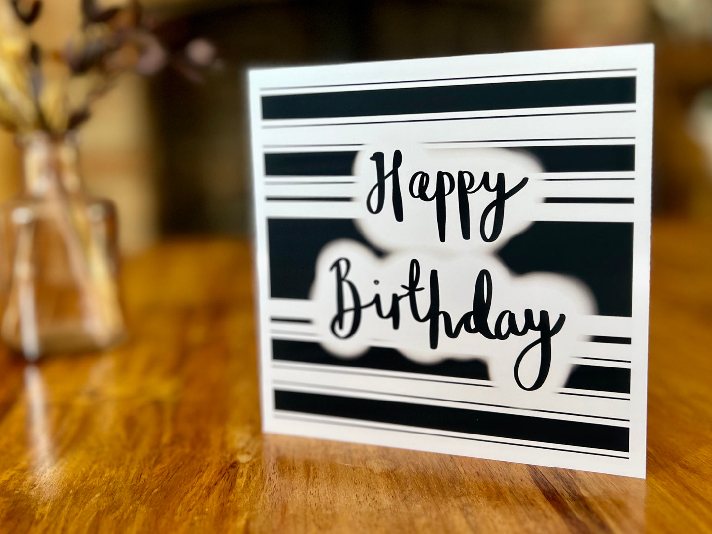 Handmade monochrome birthday card displayed upright on a wooden tabletop, featuring a black and white horizontal striped background with 'Happy Birthday' in cursive script, shown as a lifestyle product display.