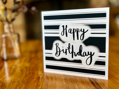 Handmade monochrome birthday card displayed upright on a wooden tabletop, featuring a black and white horizontal striped background with 'Happy Birthday' in cursive script, shown as a lifestyle product display.