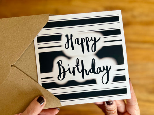 Hands holding a handmade monochrome birthday card being removed from a kraft envelope. The card features a black and white hooped background with the words 'Happy Birthday' in a cursive style, inspired by classic sports kit designs.