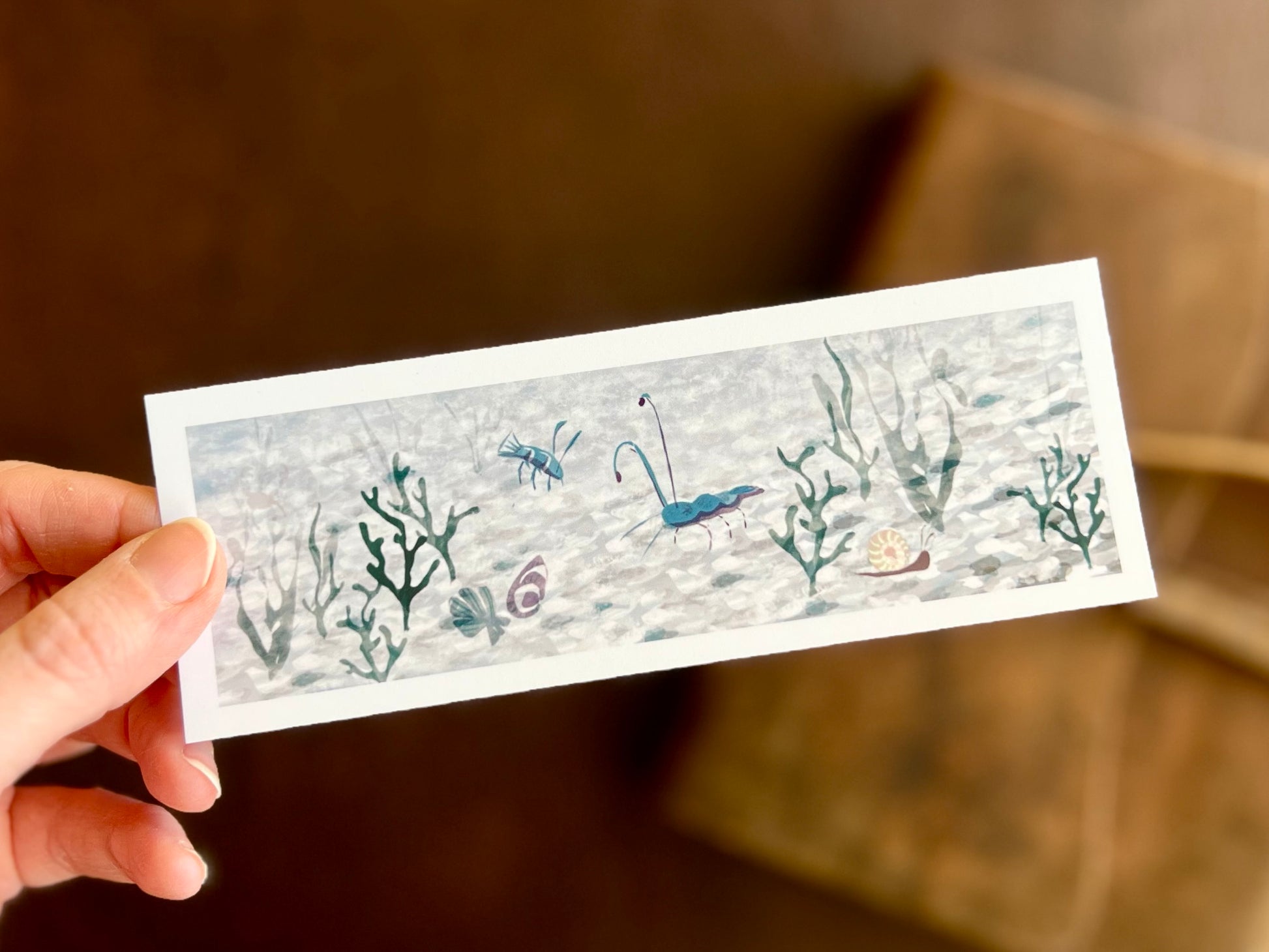Hand holding a handcrafted ocean bookmark featuring a blue prawn and blue lobster swimming in a crystal clear ocean with vibrant underwater illustration.