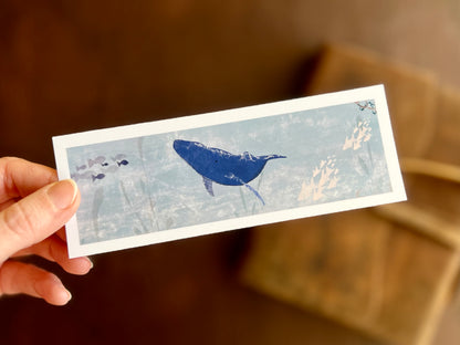 Hand holding a handcrafted blue whale bookmark featuring an original ocean illustration with a white border and matte finish.