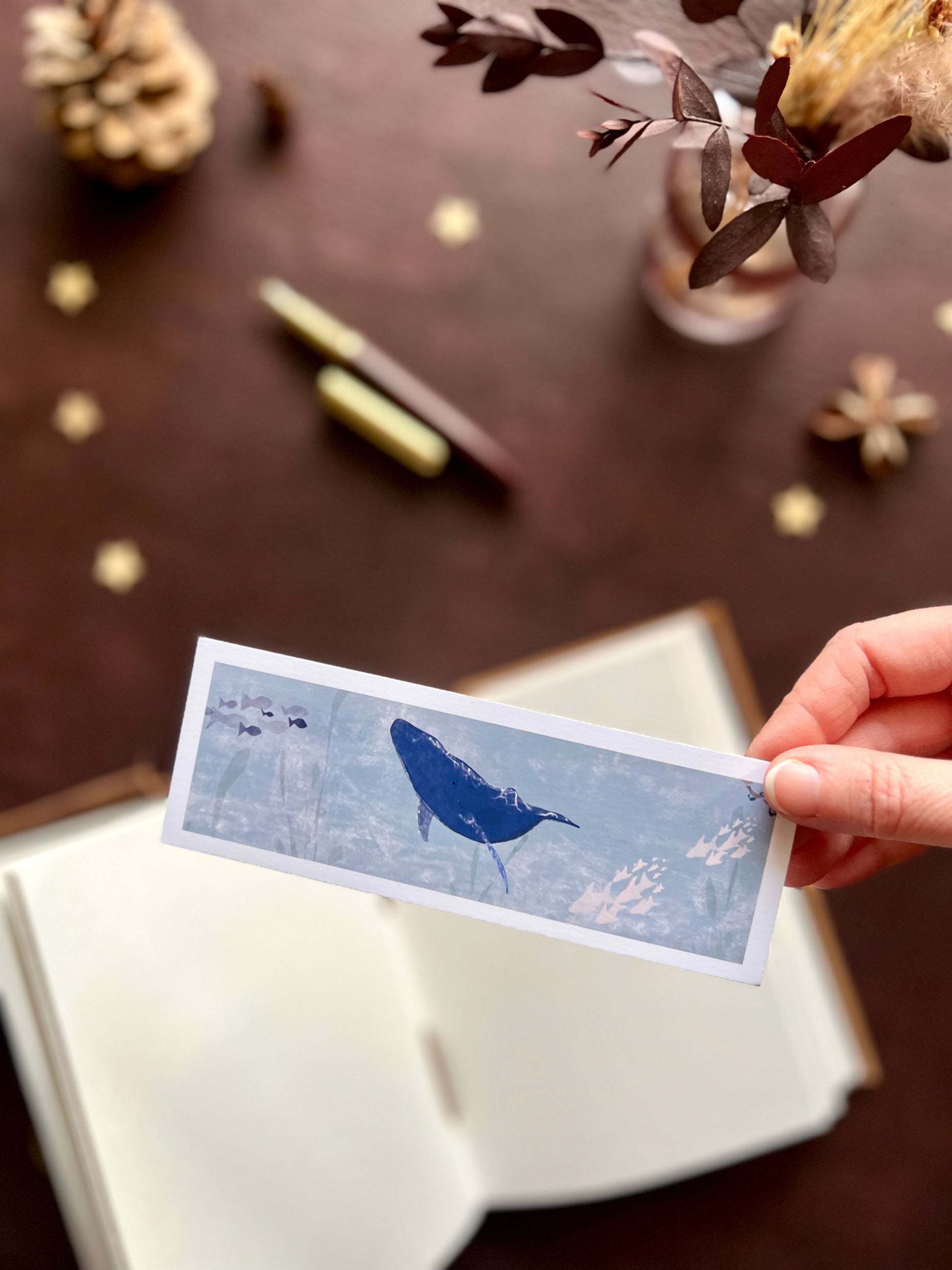 Hand holding a handcrafted blue whale bookmark with white border, styled in a nature-inspired desk scene with out of focus pine cones and flowers in the background.