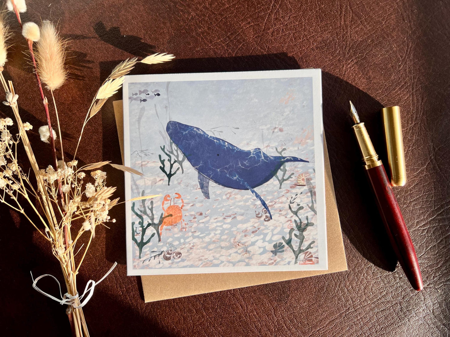 Flat lay image of a handmade greeting card featuring a blue whale and small orange crab in a pale blue ocean scene with seaweed and white pebbles, shown laid flat on top of its kraft envelope on a brown leatherette tabletop. A fountain pen and dried flowers are placed to the left and right as styling props.