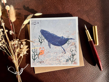 Flat lay image of a handmade greeting card featuring a blue whale and small orange crab in a pale blue ocean scene with seaweed and white pebbles, shown laid flat on top of its kraft envelope on a brown leatherette tabletop. A fountain pen and dried flowers are placed to the left and right as styling props.