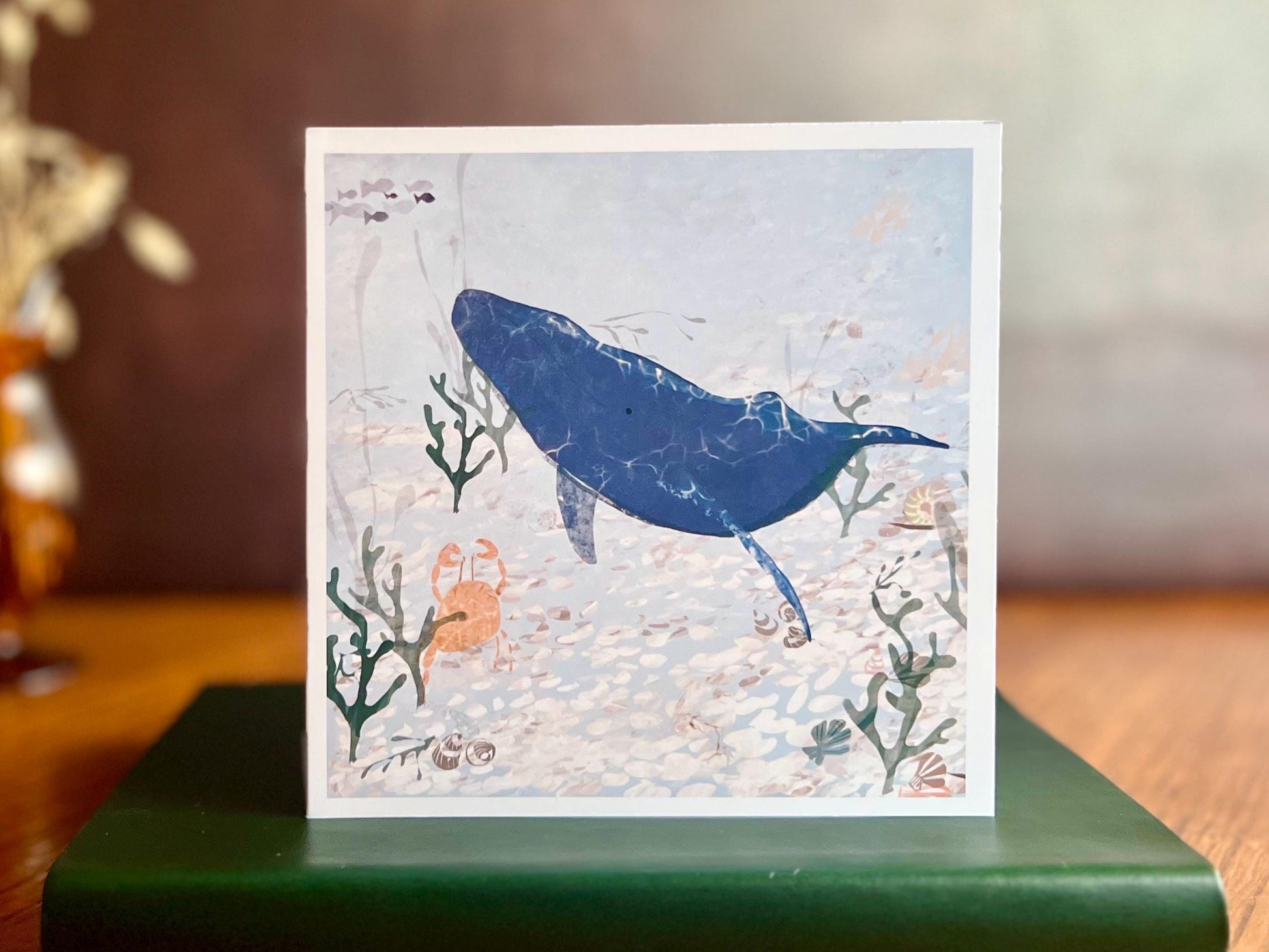 Handmade greeting card featuring a blue whale and small orange crab in a pale blue ocean scene with seaweed and white pebbles, displayed standing upright on a green leatherette book placed on a wooden tabletop in a styled lifestyle setting.