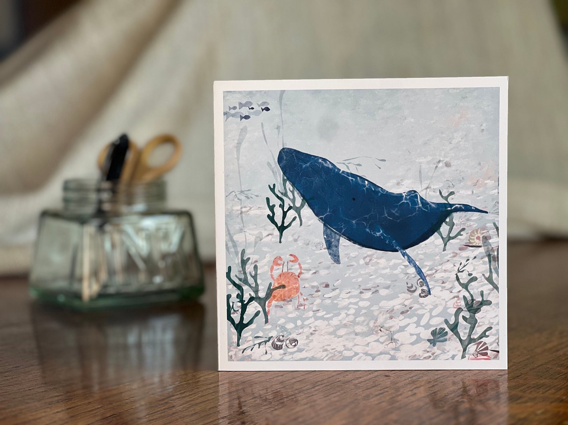 Handmade greeting card featuring a blue whale and small orange crab illustrated in a pale blue ocean with seaweed and white pebbles, shown standing upright on a tabletop with a softly blurred background for a lifestyle product display.