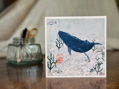Handmade greeting card featuring a blue whale and small orange crab illustrated in a pale blue ocean with seaweed and white pebbles, shown standing upright on a tabletop with a softly blurred background for a lifestyle product display.