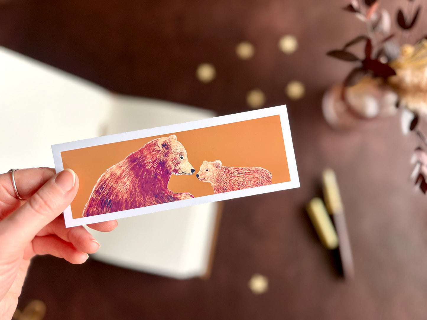 Hand holding a brown bears bookmark styled with a fountain pen and open sketchbook in a cosy autumn inspired dark academia desk scene.