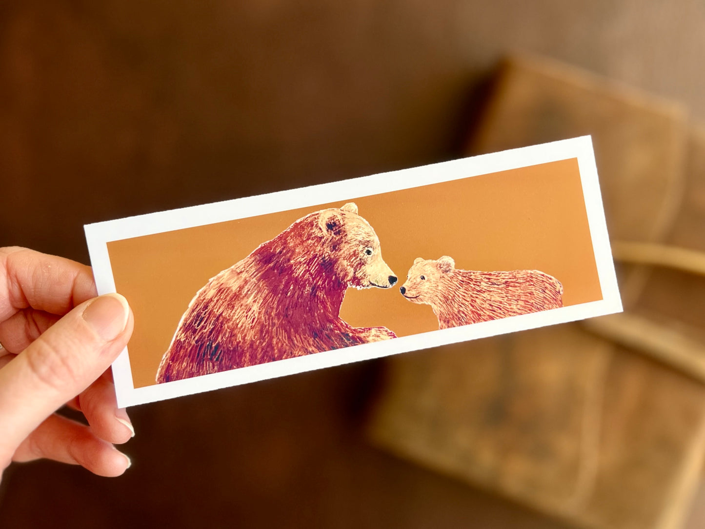 Hand holding a handcrafted brown bears bookmark featuring a bear and cub illustration on a burnt orange background.