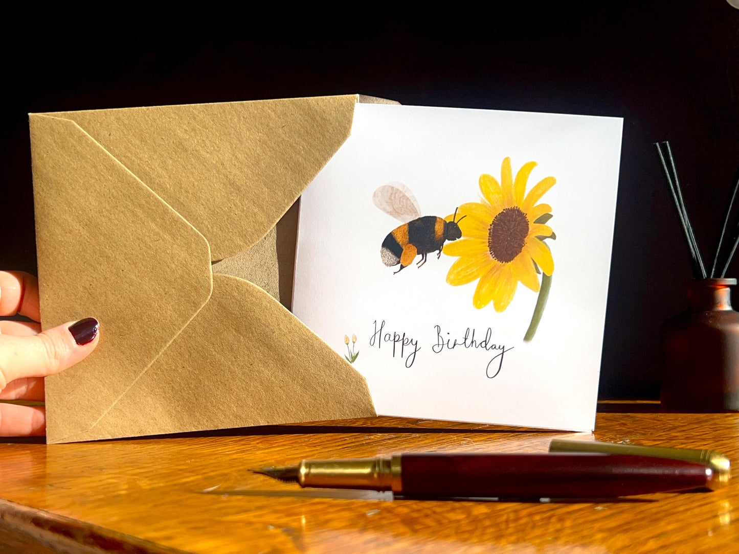 Hands holding a kraft envelope with a handmade birthday card being removed, shown upright towards the camera on a desk with a dark background. The card features an illustrated bumblebee flying towards a yellow sunflower on a white background, with a fountain pen in the foreground.