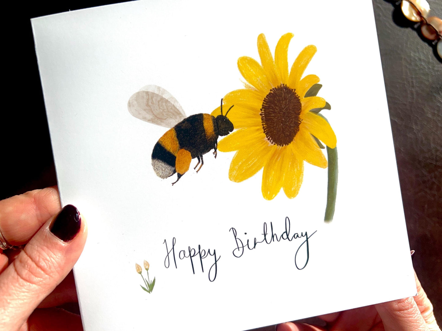 Close-up lifestyle image of a handmade birthday card being held in hands, featuring an illustrated bumblebee flying towards a yellow sunflower on a white background with 'Happy Birthday' text and a customisable name area, shown as the recipient looks at the design.