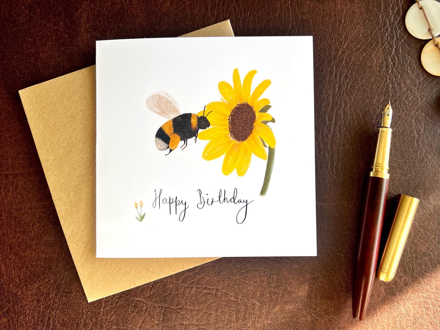 Flat lay image of a handmade birthday card featuring an illustrated bumblebee flying towards a yellow sunflower on a white background, shown laid flat on top of a kraft envelope on a brown leatherette tabletop beside a fountain pen.
