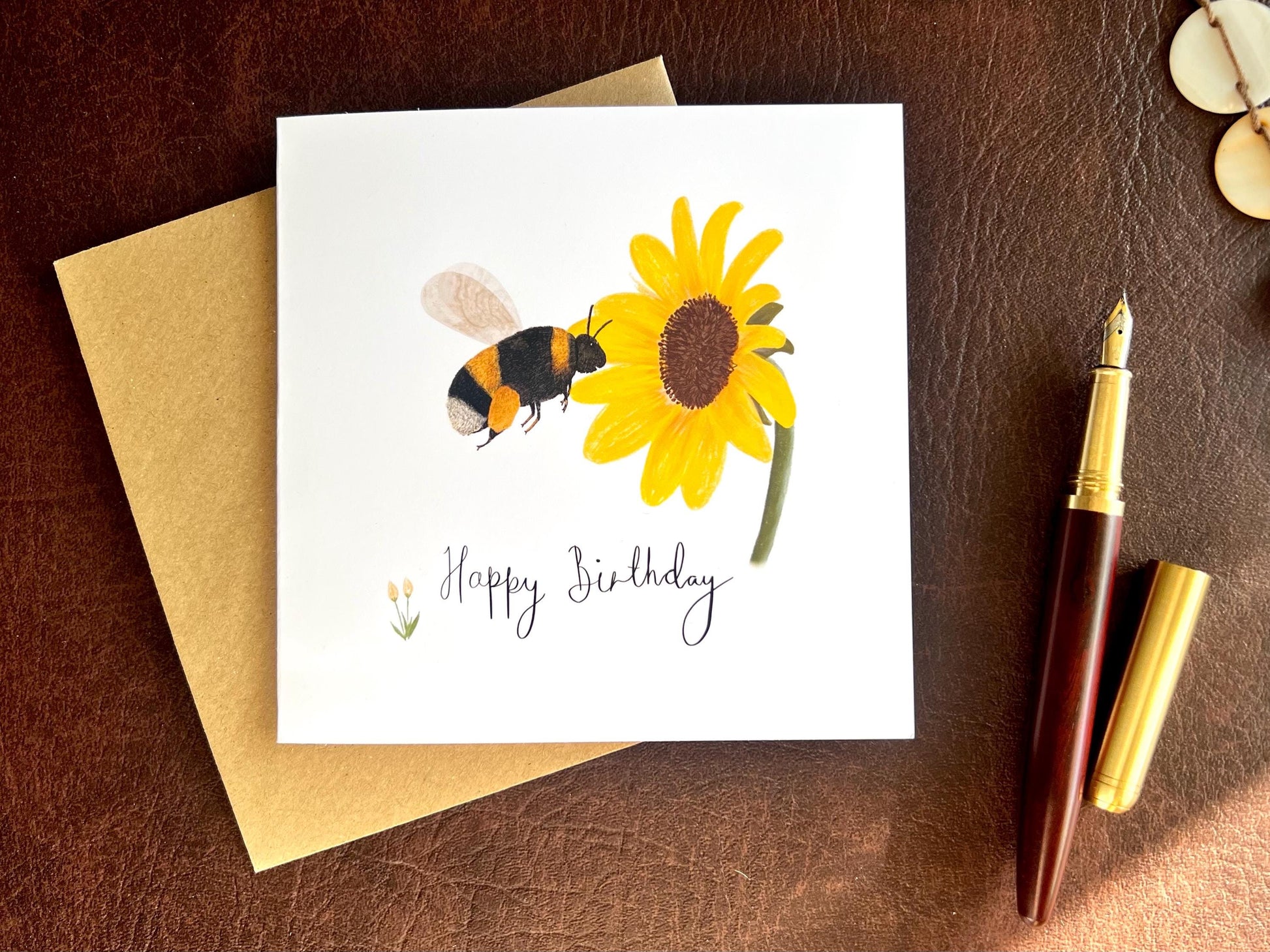 Flat lay image of a handmade birthday card featuring an illustrated bumblebee flying towards a yellow sunflower on a white background, shown laid flat on top of a kraft envelope on a brown leatherette tabletop beside a fountain pen.