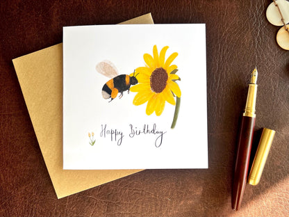 Flat lay image of a handmade birthday card featuring an illustrated bumblebee flying towards a yellow sunflower on a white background, shown laid flat on top of a kraft envelope on a brown leatherette tabletop beside a fountain pen.