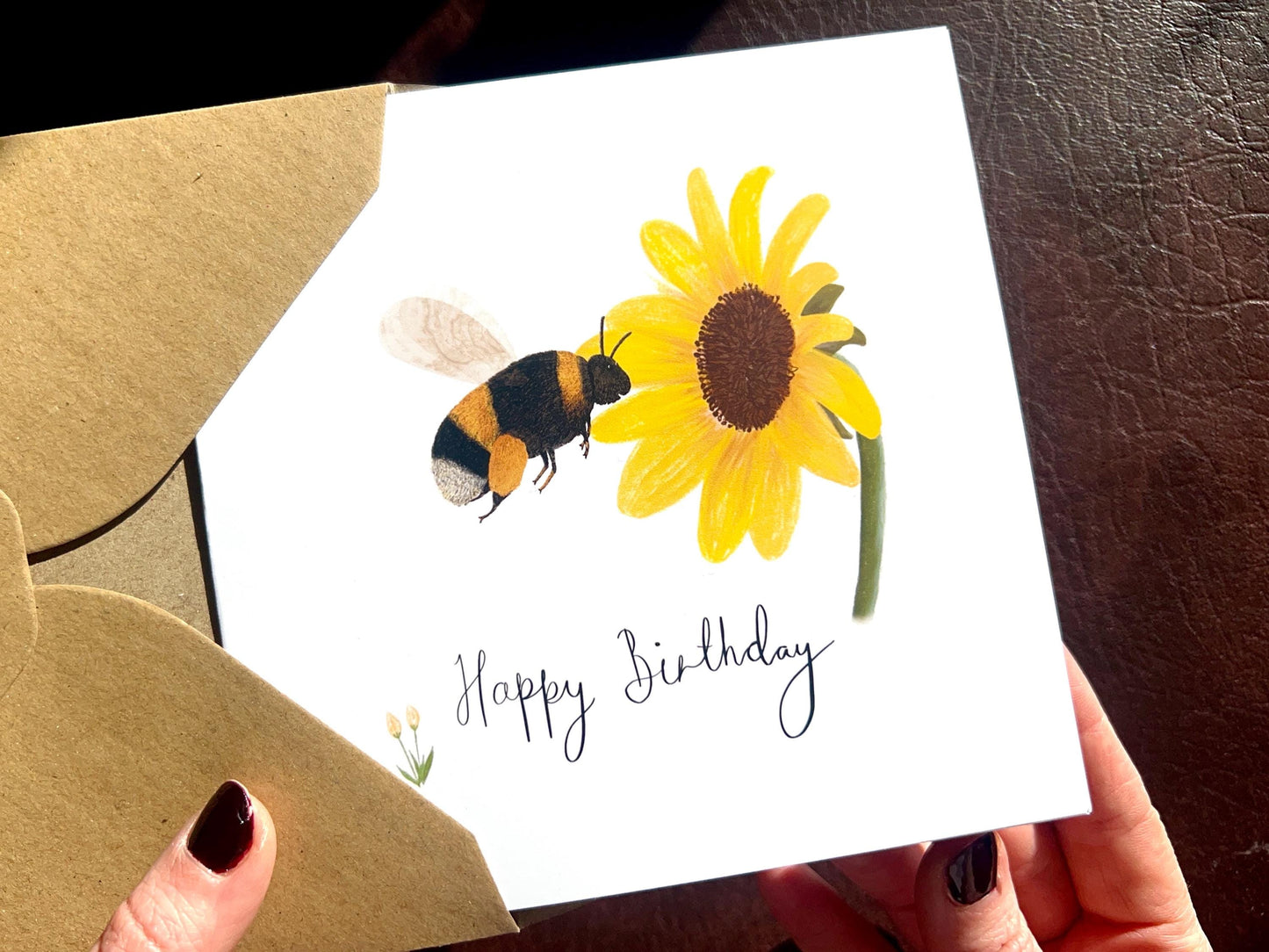 Hands holding a handmade birthday card being removed from a kraft envelope, featuring an illustrated bumblebee flying towards a yellow sunflower on a white background. The card shows 'Happy Birthday' in a swirly handwritten font without a custom name.