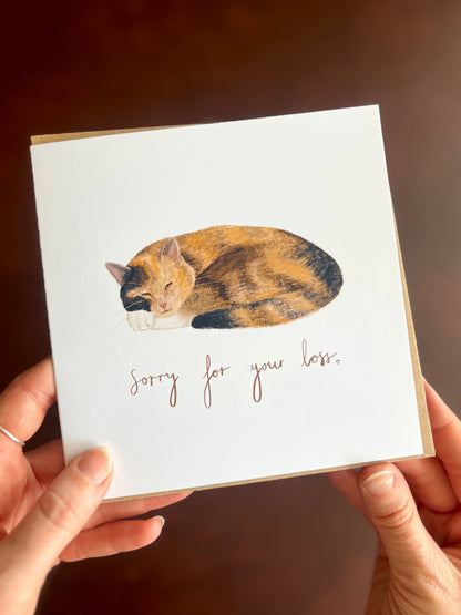 Calico cat sympathy card with “Sorry for your loss” text, featuring a peaceful sleeping ginger, black and white tortoiseshell cat illustration on a white background, held in hands with recycled kraft envelope visible.