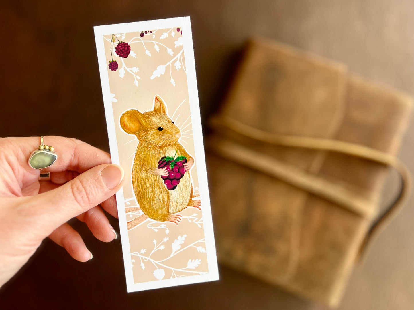 Hand holding a handcrafted countryside mouse bookmark featuring a mouse holding a blackberry on a pinky peach background with white floral detailing.