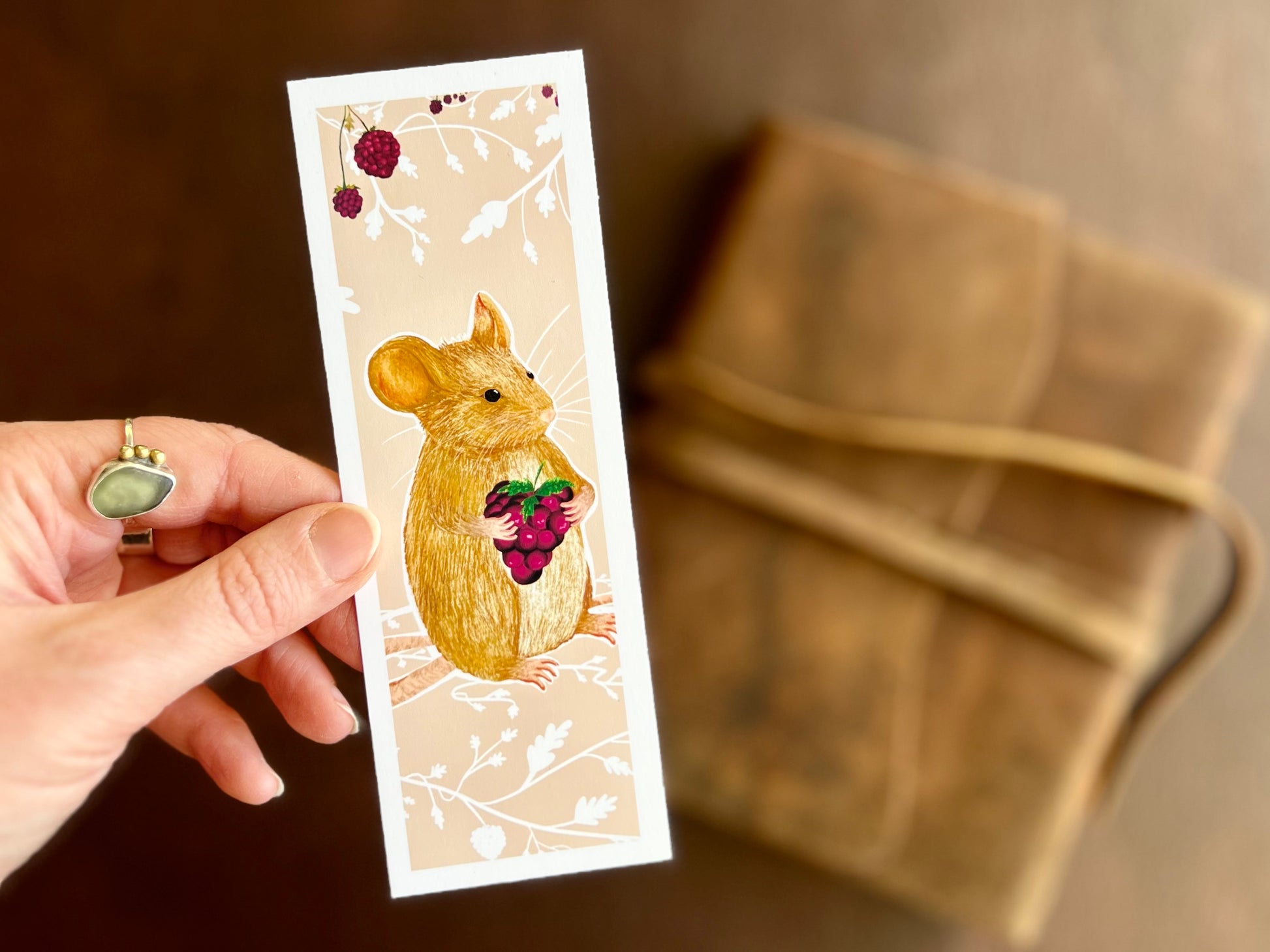 Hand holding a handcrafted countryside mouse bookmark featuring a mouse holding a blackberry on a pinky peach background with white floral detailing.
