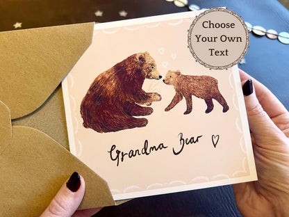 Handmade greeting card being removed from a kraft envelope, featuring a pale pink background with an illustration of a brown bear and cub. The card reads 'Grandma Bear' in a handwritten style font and includes a 'Choose Your Own Text' option for personalised wording such as grandma, gran, granny, nan or nanna, suitable for birthdays or Mother’s Day.
