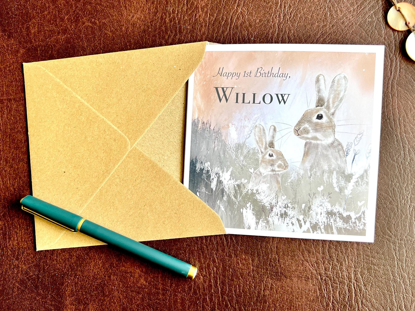 Flat lay image of a handmade customisable rabbit birthday card with a silvery grey floral illustration and pale pink evening sky design, shown partially inserted into a kraft envelope to demonstrate presentation and packaging.