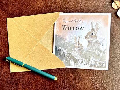 Flat lay image of a handmade customisable rabbit birthday card with a silvery grey floral illustration and pale pink evening sky design, shown partially inserted into a kraft envelope to demonstrate presentation and packaging.