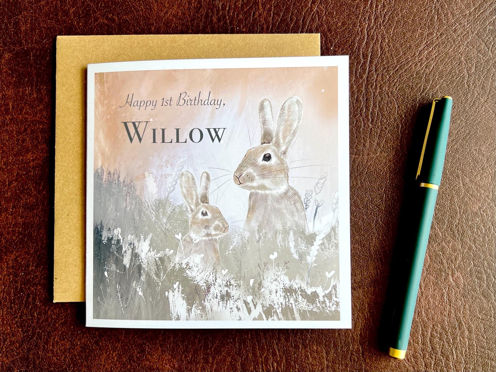 Flat lay image of a handmade customisable rabbit birthday card with a silvery grey floral illustration and pale pink evening sky background, shown laid on a brown leatherette tabletop with the kraft envelope underneath and a pen placed to the right.
