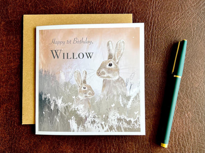 Flat lay image of a handmade customisable rabbit birthday card with a silvery grey floral illustration and pale pink evening sky background, shown laid on a brown leatherette tabletop with the kraft envelope underneath and a pen placed to the right.