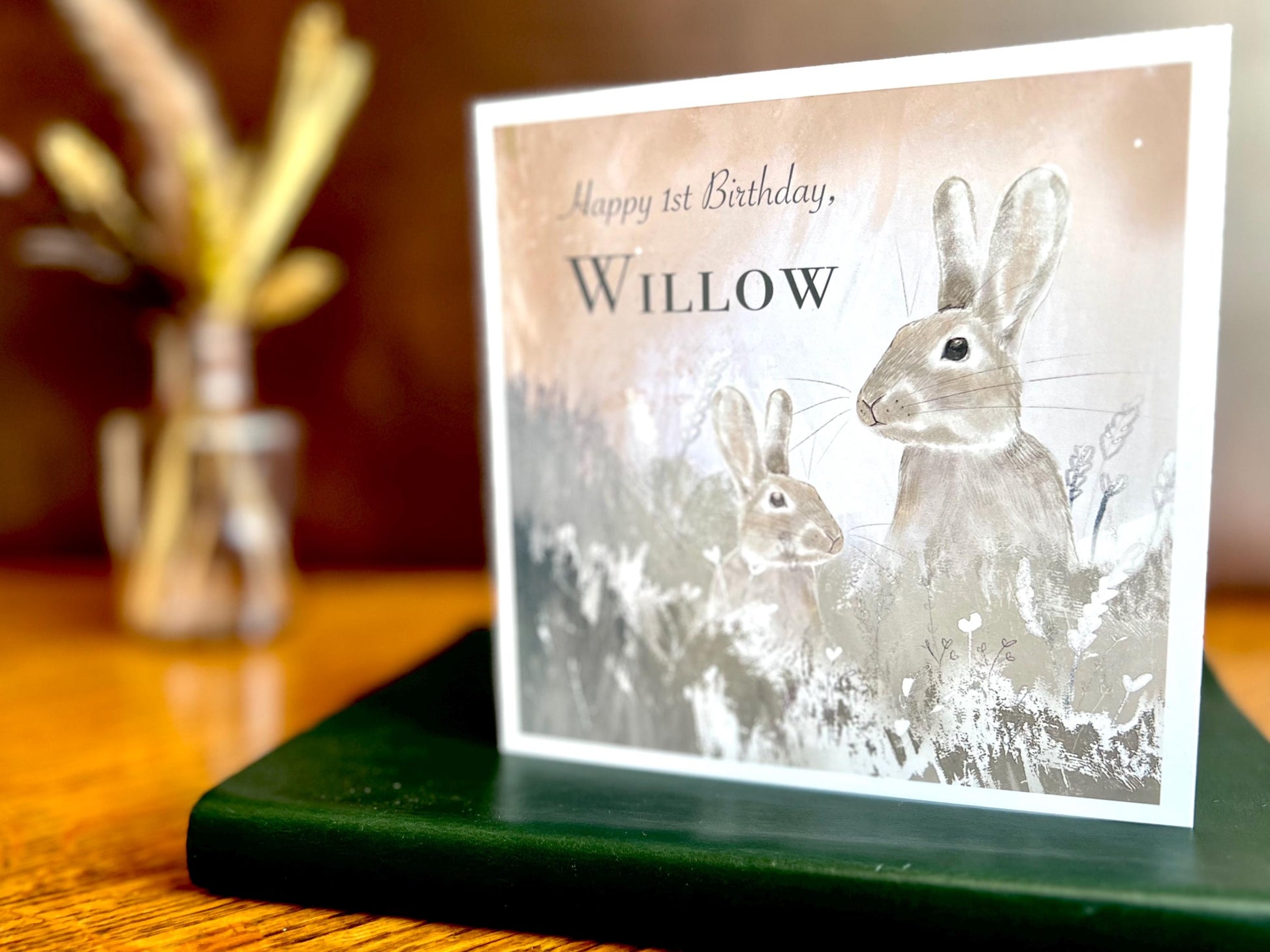 Lifestyle image of a handmade rabbit birthday card with a silvery grey floral illustration and pale pink evening sky background, displayed standing on a green leatherette book on a wooden tabletop to showcase the design.