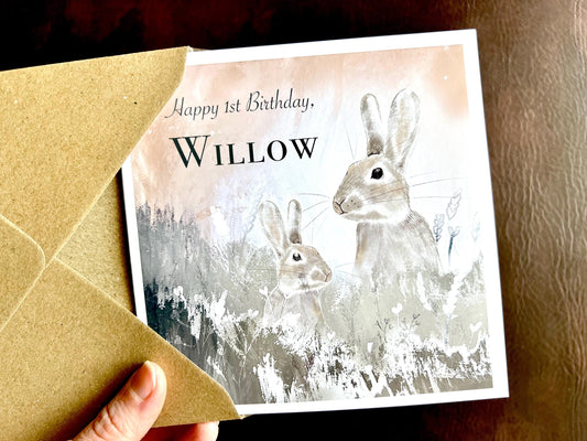 Hand holding a handmade greeting card being removed from a kraft envelope, featuring an illustration of two rabbits among flowers in silvery grey tones against a pale pink and white evening sky. The card includes customisable text for age and name, shown as 'Happy [Age] Birthday [Custom Name]'.