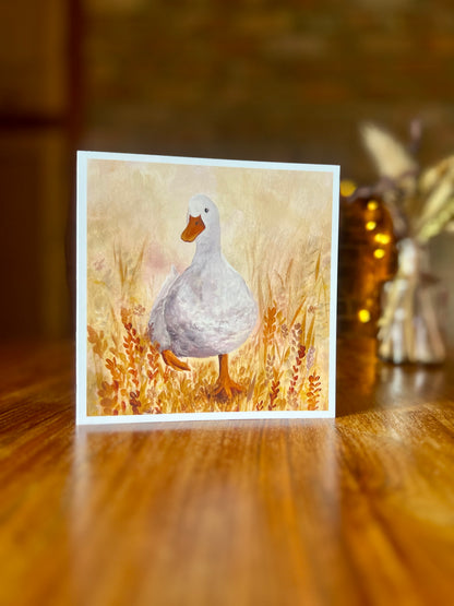 Artist-designed greeting card featuring a print of the original oil painting Duck in the Meadow, showing a characterful white duck waddling through a colourful meadow of yellow and orange flowers in a painterly style with a white border.