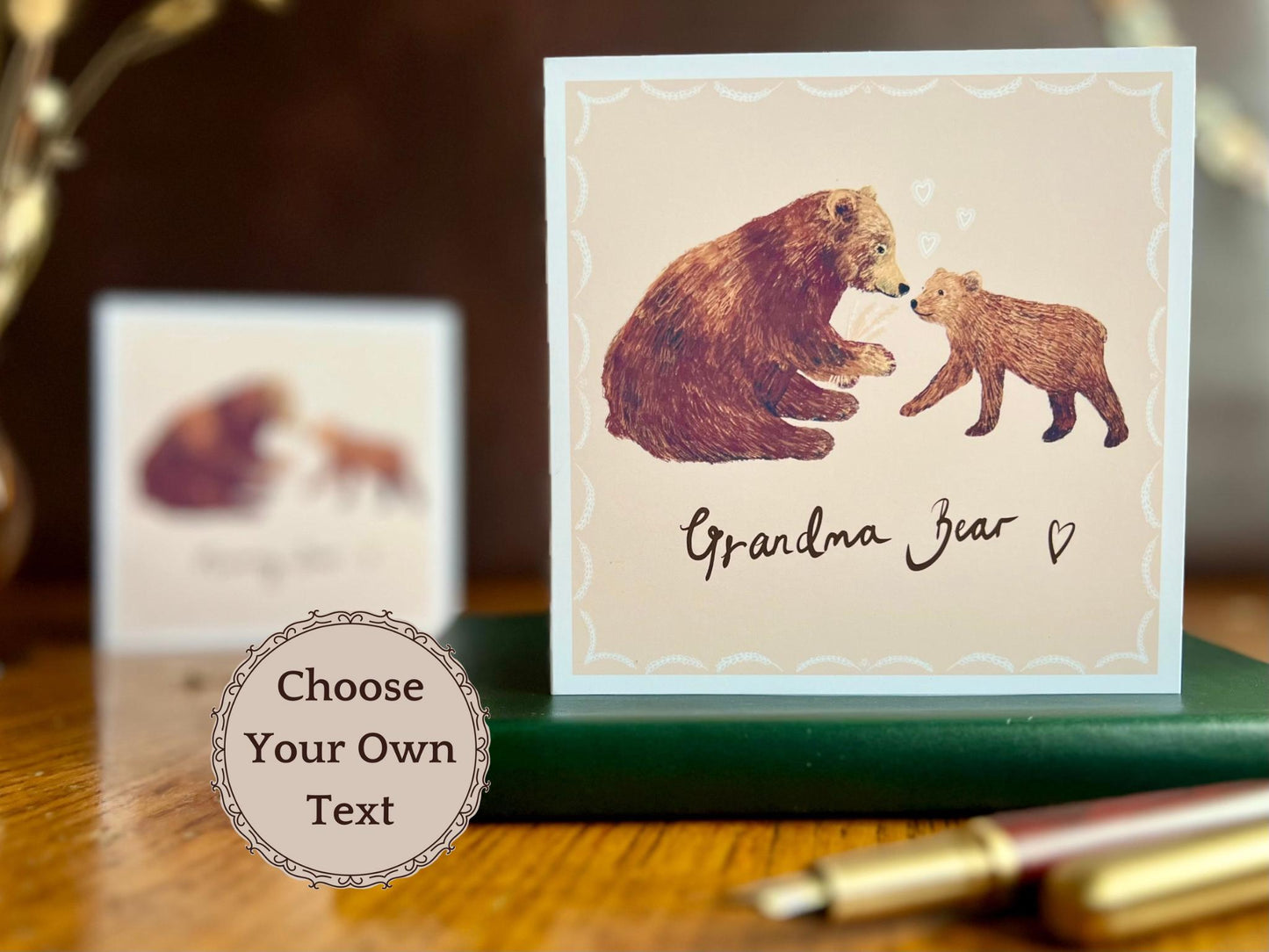 Lifestyle image of a handmade greeting card featuring a brown bear and cub illustration titled 'Grandma Bear', displayed on a leatherette book on a tabletop with a fountain pen in the foreground. The background shows a blurred version of the same card with different wording, and a text overlay reading 'Choose Your Own Text', highlighting its customisable design for family names such as grandma, gran or nan.