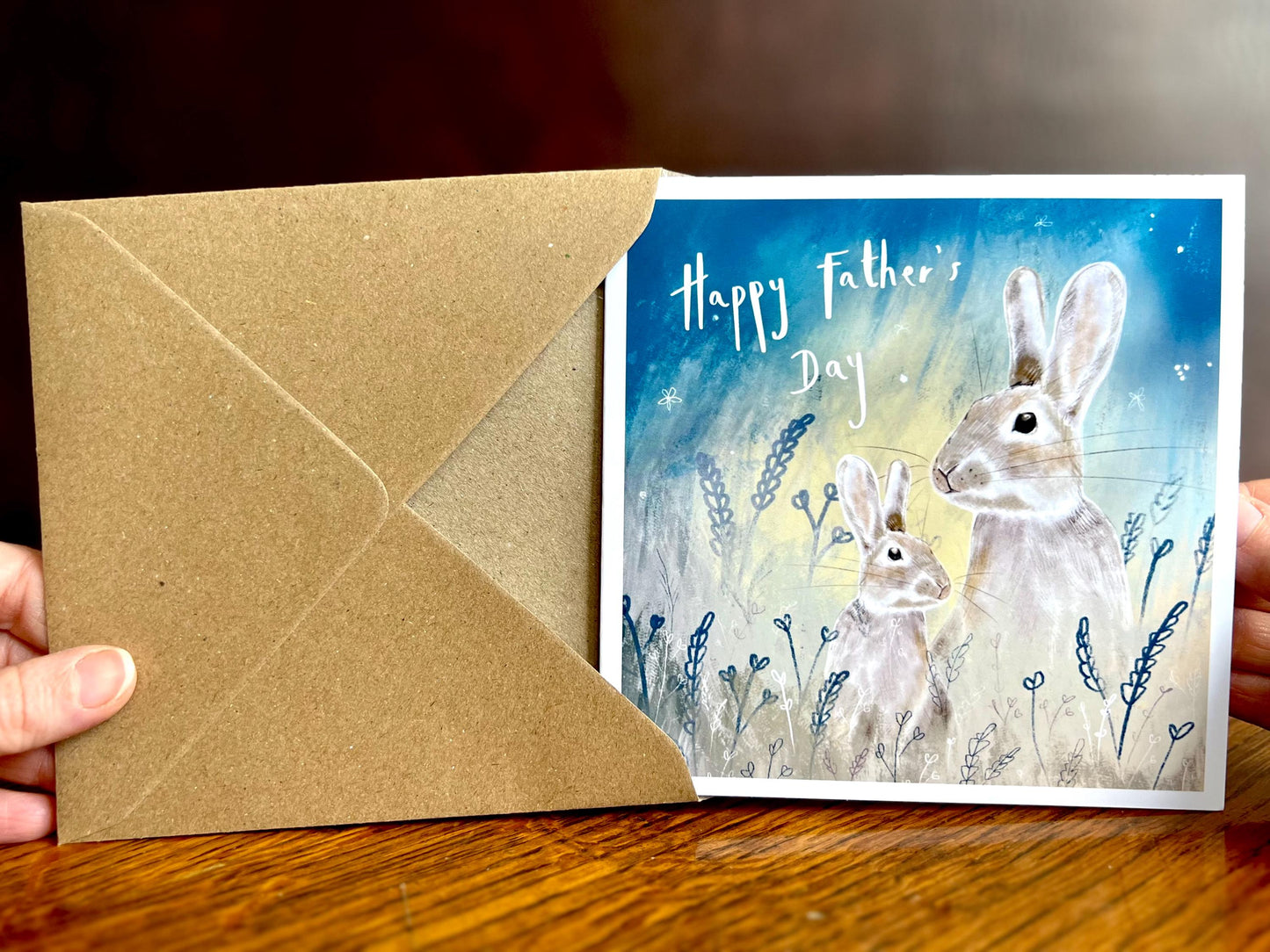 Handcrafted Father’s Day greeting card and recycled fleck kraft envelope being held in hands to showcase texture and detail. The card features an illustrated father rabbit and young rabbit under a starry night sky with foliage and “Happy Father’s Day” text on the front, highlighting premium illustration quality.