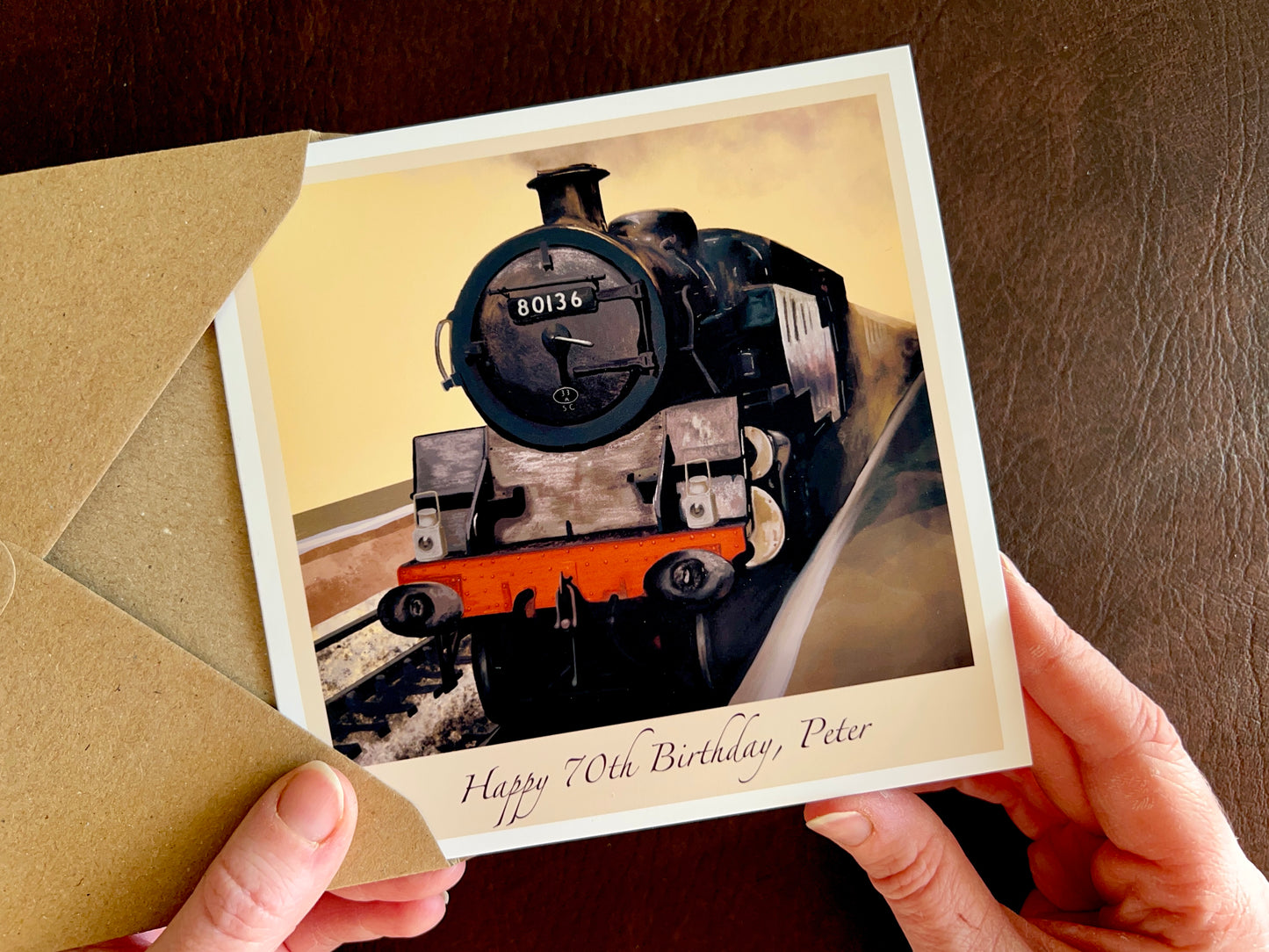 Hands holding a handcrafted greeting card featuring an illustrated steam train, engine 80136 from the North Yorkshire Moors Railway, with personalised text reading “Happy 70th Birthday, [Name]”. Customisable birthday card with illustrated design and recycled kraft envelope.