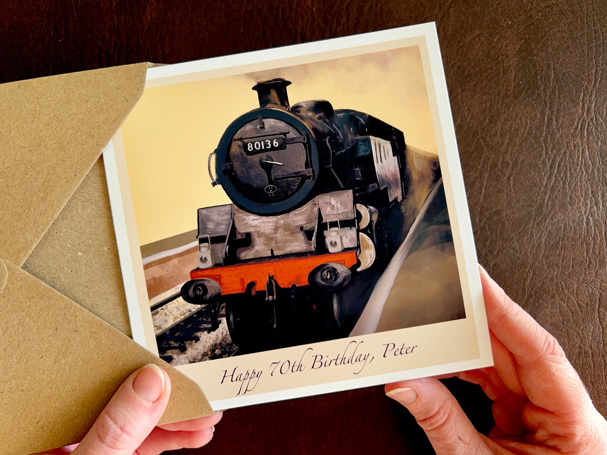 Hands holding a handcrafted greeting card featuring an illustrated steam train, engine 80136 from the North Yorkshire Moors Railway, with personalised text reading “Happy 70th Birthday, [Name]”. Customisable birthday card with illustrated design and recycled kraft envelope.