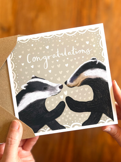 Hands gently opening an envelope to reveal a hand-crafted engagement congratulations card featuring a picture-book style badger illustration from a recycled kraft envelope, rustic woodland themed design on soft sage green background, gender neutral engagement card for engaged couples including Mr & Mr, Mrs & Mrs or Mr & Mrs, also suitable as a thoughtful congratulations gift for son and daughter in law or family celebrations