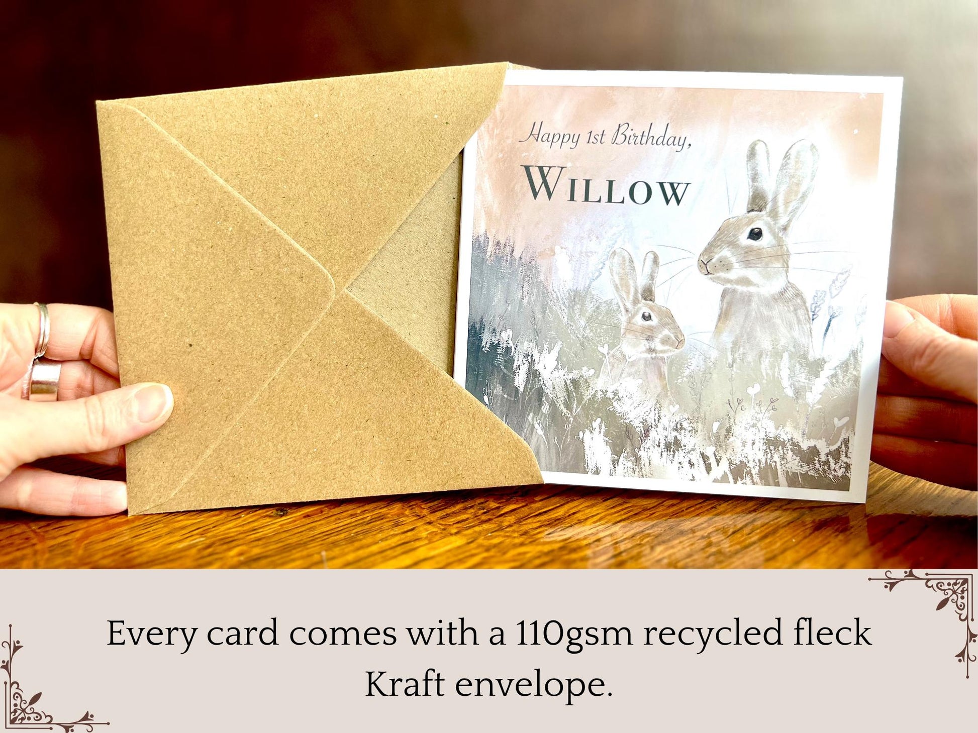 Hand holding a handmade customisable rabbit birthday card being removed from a kraft envelope, shown upright towards the camera. The card features a silvery grey floral illustration with a pale pink evening sky background, and text highlighting that every card comes with a 110gsm recycled fleck Kraft envelope.