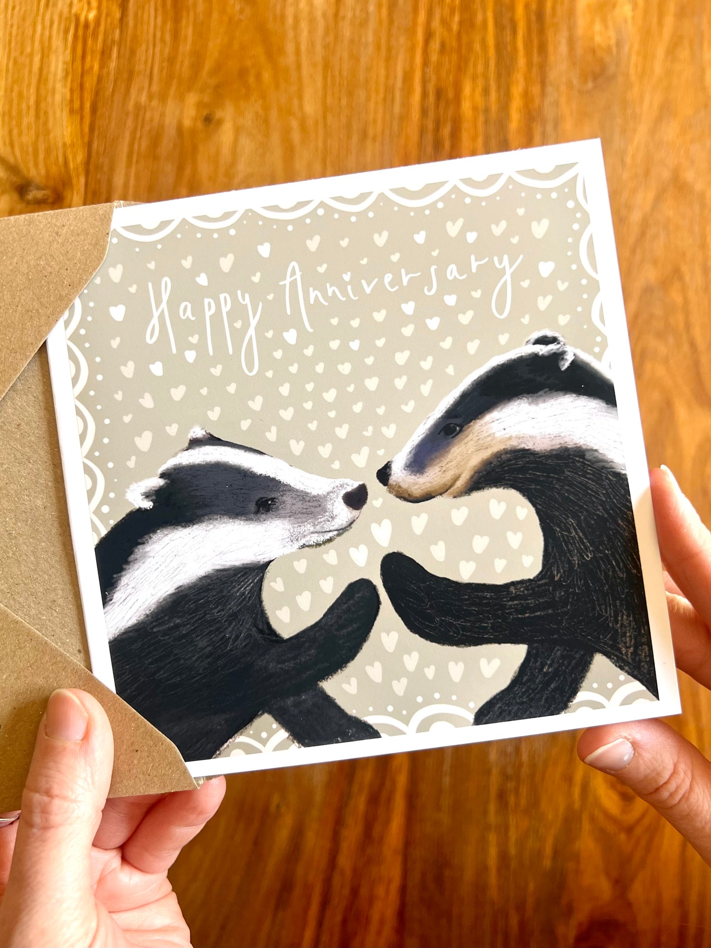 Hands holding a Happy Anniversary greeting card featuring two black and white woodland badgers touching paw to paw on a pale sage green background with hearts, a premium high quality artist-designed art card.