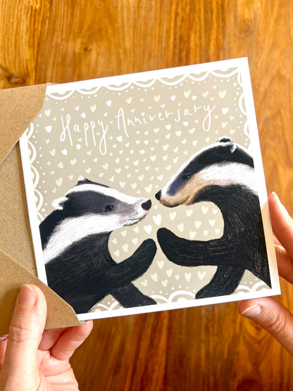 Hands holding a Happy Anniversary greeting card featuring two black and white woodland badgers touching paw to paw on a pale sage green background with hearts, a premium high quality artist-designed art card.