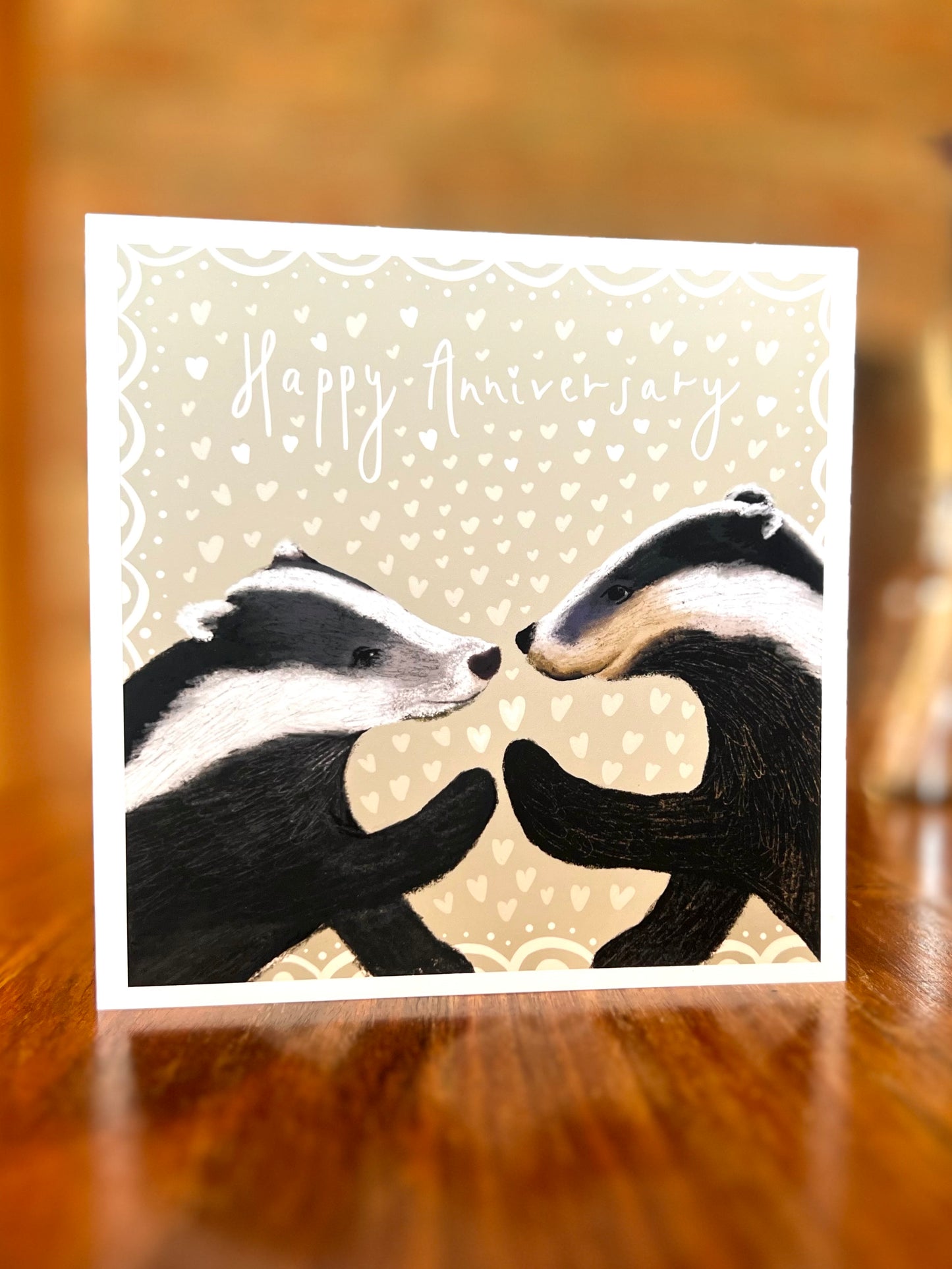 Happy Anniversary greeting card standing on a rustic tabletop in a farmhouse setting, featuring two black and white woodland badgers touching paw to paw on a pale sage green background with hearts, a premium high quality artist-designed art card.