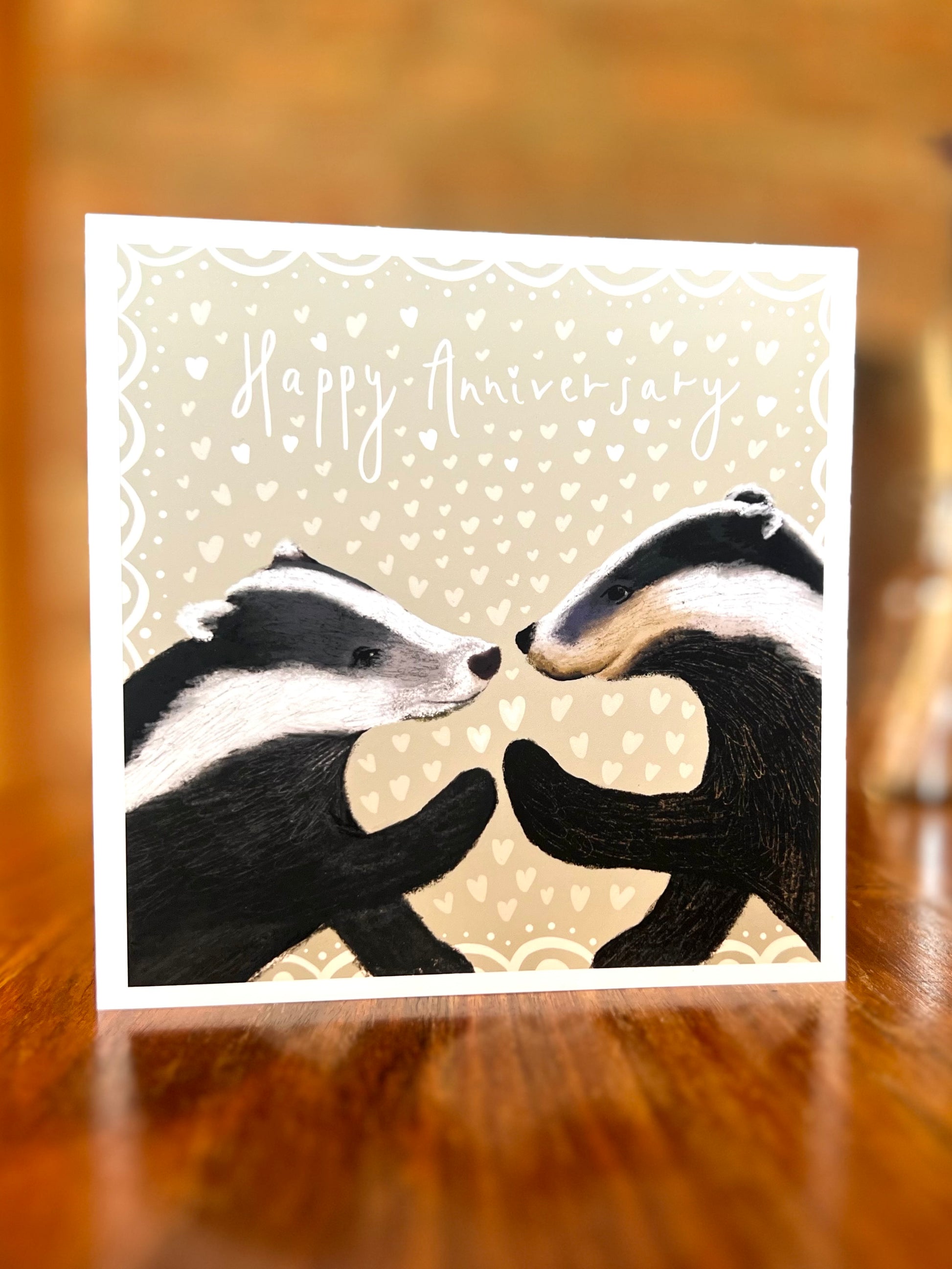 Happy Anniversary greeting card standing on a rustic tabletop in a farmhouse setting, featuring two black and white woodland badgers touching paw to paw on a pale sage green background with hearts, a premium high quality artist-designed art card.