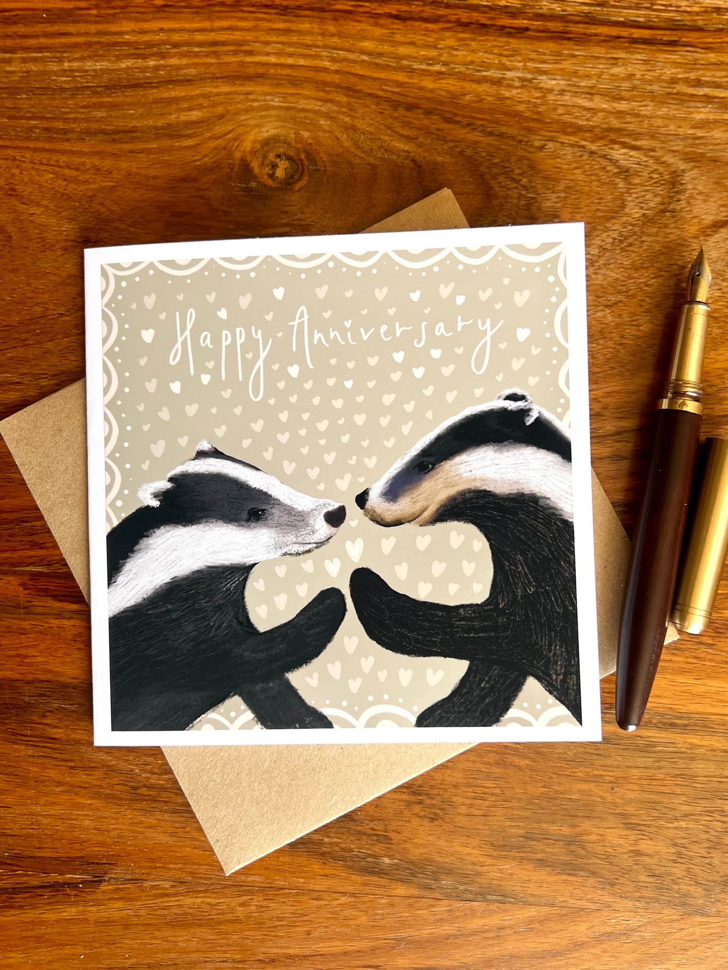 Happy Anniversary greeting card laid flat on a wooden tabletop on top of a recycled kraft envelope, featuring two black and white woodland badgers touching paw to paw on a pale sage green background with hearts, alongside a wooden fountain pen, premium high quality artist-designed art card.