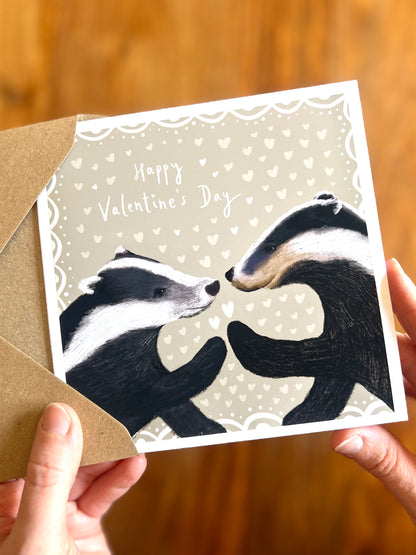 Hands holding a Valentine’s Day greeting card featuring two black and white woodland badgers touching paw to paw on a pale sage green background with subtle white hearts and the text Happy Valentine’s Day.