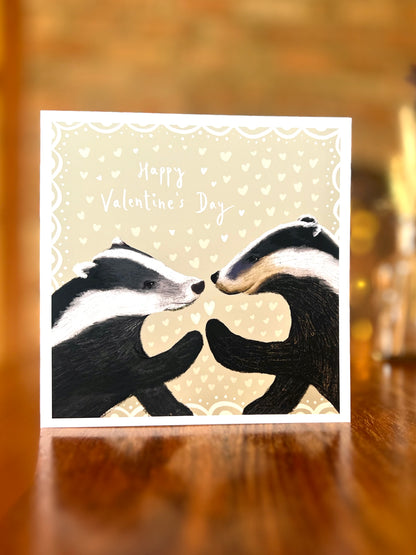 Happy Valentine’s Day greeting card standing on a rustic wooden tabletop, featuring two black and white woodland badgers touching paw to paw on a pale sage green background with subtle white hearts.