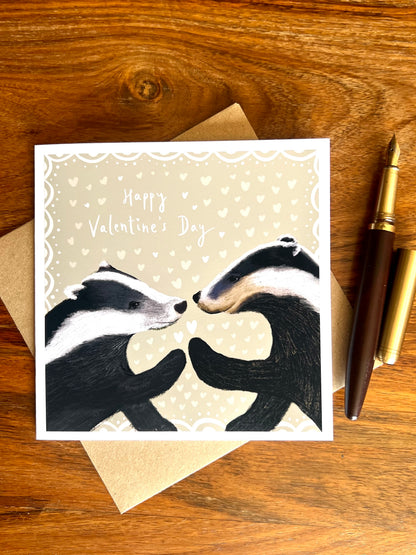 Happy Valentine’s Day greeting card laid flat on a rustic tabletop on top of a recycled fleck kraft envelope, featuring two black and white woodland badgers touching paw to paw, with a wooden fountain pen beside the card.