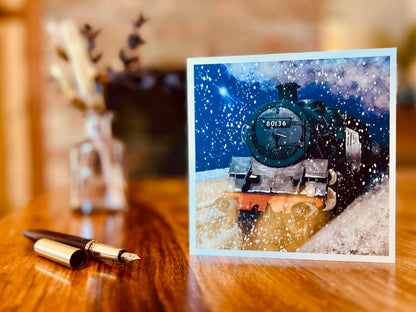Artisan Christmas card featuring steam train 80136 on a snowy winter night with falling snow and glowing headlights, displayed upright on a wooden tabletop. A wooden fountain pen lies next to the card and a blurred vase sits in the background.