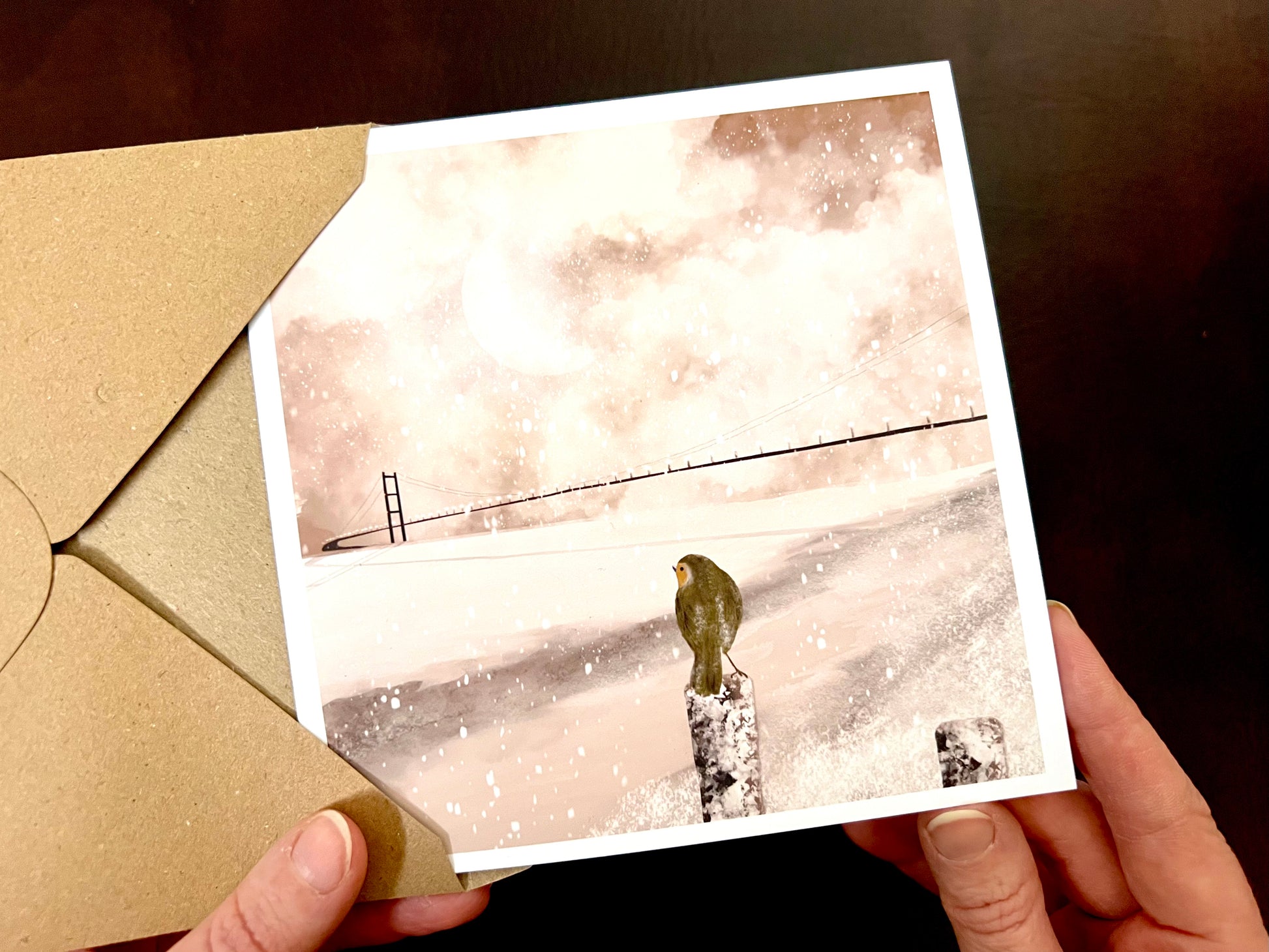 Premium winter greeting card featuring an illustrated snowy scene of the Humber Bridge linking Hessle near Hull in East Yorkshire to Barton in North Lincolnshire. A small robin is perched on a post overlooking the view, with soft snow falling and sunlight breaking through pink and white winter clouds reflecting on the water. The square card has a white border and is ideal as a Christmas card or seasonal greeting for those in Hull, East Yorkshire and North Lincolnshire. Printed on thick 312gsm fine art card