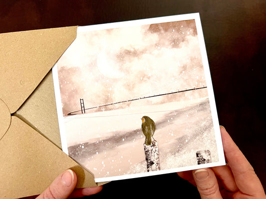 Premium winter greeting card featuring an illustrated snowy scene of the Humber Bridge linking Hessle near Hull in East Yorkshire to Barton in North Lincolnshire. A small robin is perched on a post overlooking the view, with soft snow falling and sunlight breaking through pink and white winter clouds reflecting on the water. The square card has a white border and is ideal as a Christmas card or seasonal greeting for those in Hull, East Yorkshire and North Lincolnshire. Printed on thick 312gsm fine art card