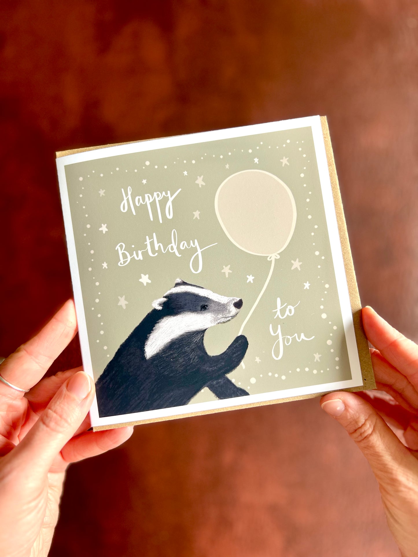 Artist designed woodland animal birthday card held by hands over rustic background featuring cute illustrated badger and handwritten lettering