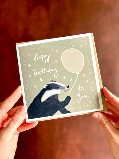 Artist designed woodland animal birthday card held by hands over rustic background featuring cute illustrated badger and handwritten lettering