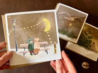 Premium Christmas card from a set of three woodland designs, featuring two mice wearing winter hats and coats sitting together on a bench in a snowy night scene beneath a fairy-lit branch. The card is shown in close-up being held in two hands, with the other two designs softly out of focus in the background. Ideal festive greeting card for animal lovers and rustic Christmas card sets. Printed on thick 312gsm fine art card by a UK independent artist.